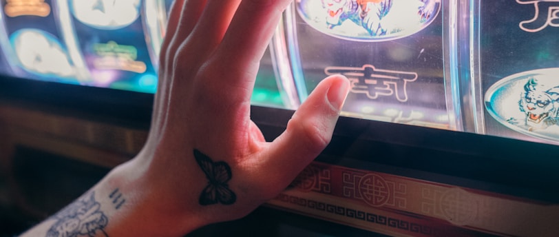 A hand with tattoos reaches towards a brightly lit slot machine, featuring various colorful symbols including a tiger. The image captures the moment of anticipation as the person engages with the machine during a game of chance.