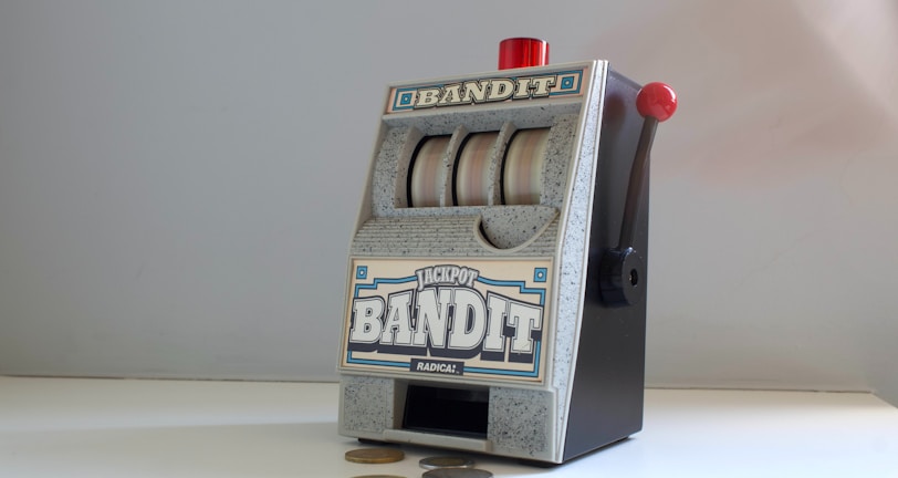 A small, vintage-style slot machine with the word 'Bandit' prominently displayed. It features a lever on the side with a red knob and three reels with a generic design. Several coins are scattered on the white surface in front of it.